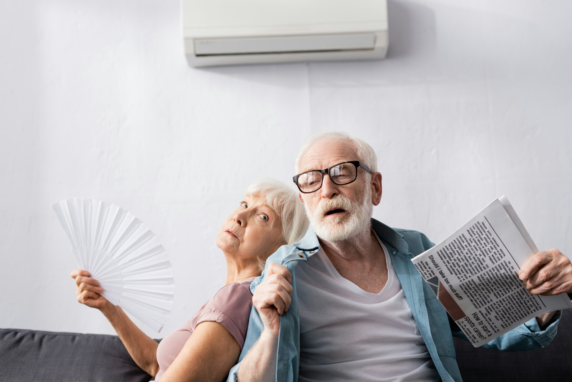 Senior man holding newspaper near tired wife with fan under air conditioner in living room